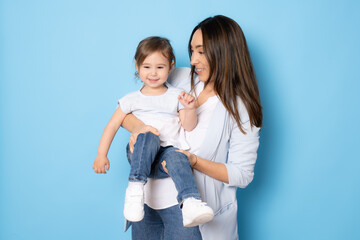 Photo of pretty young mommy hugging little daughter standing isolated over blue background. Motherhood concept.
