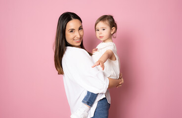 Photo of pretty young mommy hugging little daughter standing isolated over pink background. Motherhood concept.