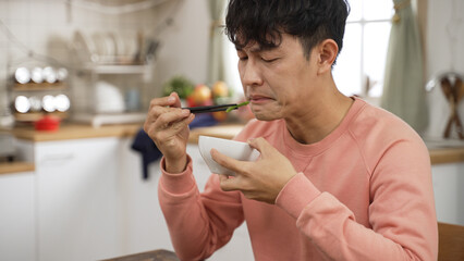 asian Japanese man grimacing face while trying a piece of vegetable in dining room at home. he...