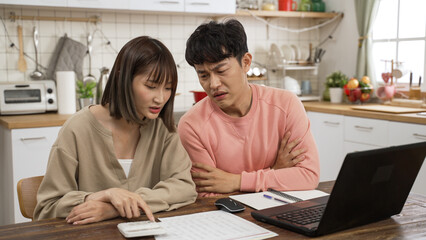 stressed Asian young couple making budget plan for mortgage loans bill. the husband folds arms as his wife is showing him figure on the calculator and bills