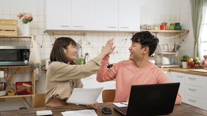 happy Asian couple high five with each other celebrating getting loan mortgage approval. they read document from bank and check information on computer