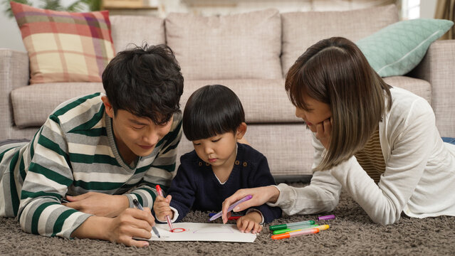 loving asian dad and mom teaching their baby boy how to paint with color pens at home. they lie on belly in the living room at leisure