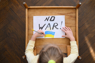Ukrainian girl kid protesting against war conflict and drawing message text No War with Ukrainian flag on the white paper. No war, stop war, russian aggression, crisis, peace, stop aggression