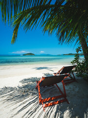 Scenic view of Koh Mak Island's peaceful clear turquoise water bay, relax chair on white sand beach with coconut palm tree foreground. Koh Kham Island view at horizon. Trat, Thailand.