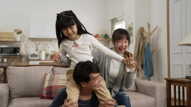 cheerful asian family of three playing together at home. the father carrying his daughter on shoulders while the mother is helping hold the girls’ hands