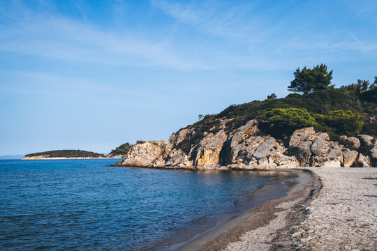View of the coast beach in the background a rocky ledge covered with green trees and bushes. Ormos Panagias, Greece.