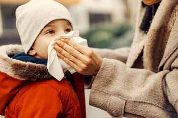 A mother blowing the nose of her little son at the park in cold weather.