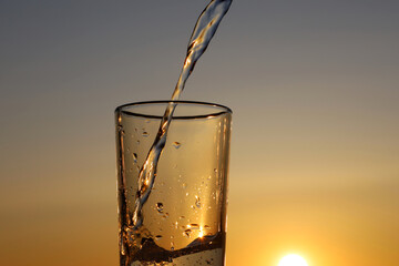 Clean water pouring into drinking glass on sunset sky background. Concept of health and freshness, thirst, water purification