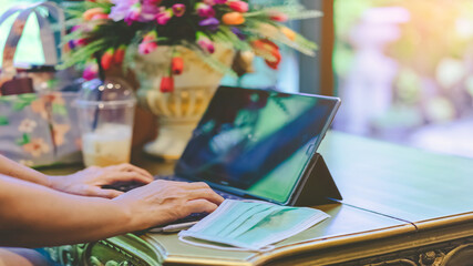 Hands of mature woman typing on laptop keyboard to work from home and take off her surgical mask put it on the table for easy breathing. Concepts of Covid-19 (Coronavirus).  Selective focus on hand.