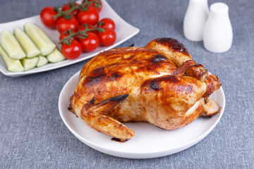 Chicken baked whole to a delicious crust. A traditional dish. In the background are cucumbers and cherry tomatoes on a white plate. Close-up, selective focus, gray background.