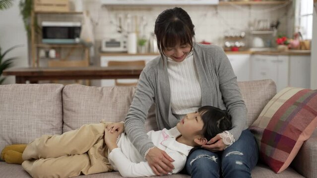 loving asian mother touching her daughter gently and talking to her while girl is lying on her laps on the sofa in the living room at home. love and family concept