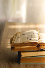 Open book and reading glasses on the table, illuminated by sunlight. Stack of vintage books in the background. Selective focus.