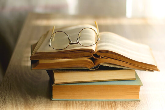 Open book and reading glasses on the table, illuminated by sunlight. Stack of vintage books in the background. Selective focus.