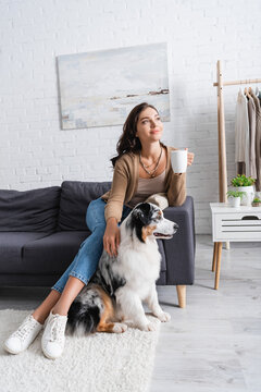 smiling young woman sitting on sofa and cuddling australian shepherd dog while holding cup of coffee.