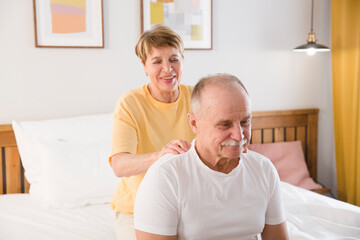 elderly woman gives a shoulder massage to her husband sitting on a bed at home. elderly couple healthy lifestyle. 