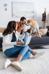 smiling young woman with cup sitting on carpet and using laptop near boyfriend cuddling akita inu dog.