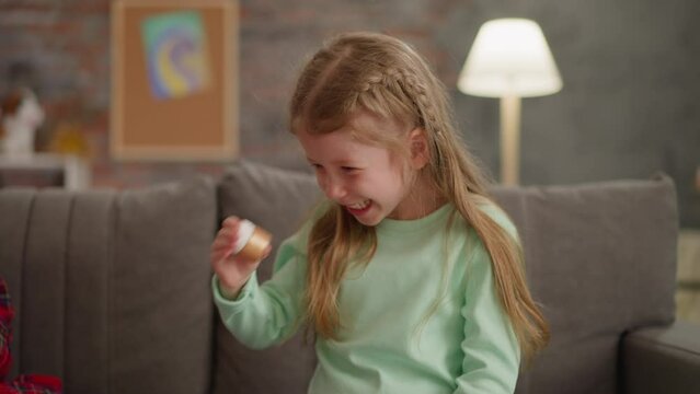 Joyful little girl shakes bottle with golden glitter on sofa