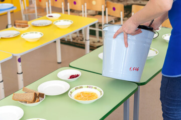 A kindergarten teacher puts soup on plates by scooping it with a ladle from a bucket.