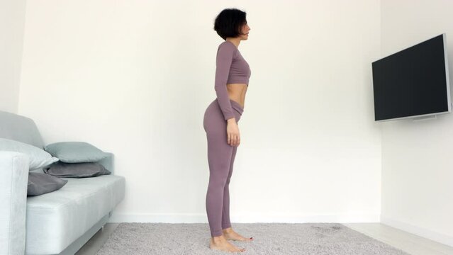 Side view young woman in violet activewear stands on carpet do butt exercises deep squat hold position improve body shape routine at home, healthy active lifestyle calories burn weight loss concept
