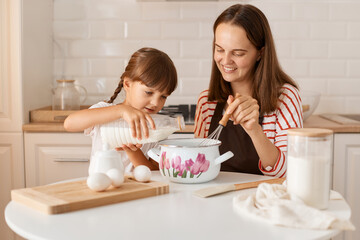 Horizontal shot of positive optimistic young adult mother and her cute little daughter baking in kitchen at home, cute kid pouring milk to pot and mom mixing dough.