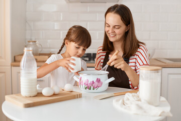 Obraz na płótnie Canvas Attractive satisfied young mother and her little daughter baking in kitchen at home, preparing homemade pastry together, expressing happiness, female kid ad sugar.