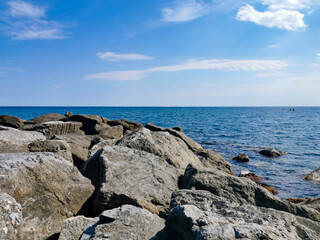 Fototapeta na wymiar Large stones on the seashore on the background a blue sky with white clouds. Large stones on the seashore on the background a blue sky with white clouds.The Ligurian Sea, Italy.