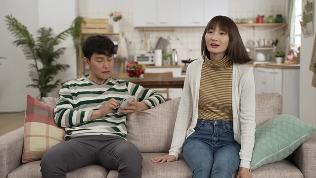 upset asian mother looking at baby son running around in living room with folded arms while the addicted father playing with mobile phone ignoring her
