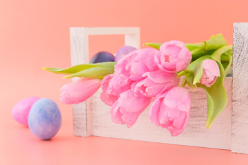A beautiful delicate top bouquet of live spring pink tulips and pink, purple, blue eggs in a light wooden box on a pink background. Easter concept. Greeting card.