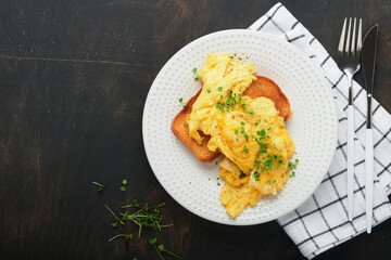 Scrambled Eggs. Fluffy and Buttery scrambled eggs on bread with microgreen radish and hollandaise sauce on white plate over dark old wooden background. Homemade breakfast or brunch meal. Top view.