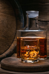 Vertical photo of whiskey in glass of ice and old barrel of old whiskey in wine cellar. Glass of bourbon whiskey.