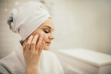 Young woman wearing white bathrobe and towel on head applying cream on face. Daily beauty routine.