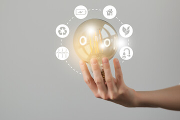 Woman`s hand holding glowing light bulb with energy resources icons. Ecological friendly and sustainable environment concept.