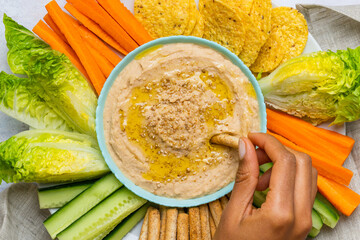 White bean hummus alongside cut vegetables. A woman's hand dipping the vegetable into the bowl.