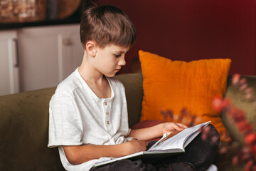 Smiling attractive teen boy sitting at home on sofa and reading book. Cheerful guy in white t-shirt is sitting at home, reading comic book. Schoolboy is reading paper book while resting home.