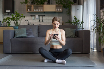 Woman doing yoga at home sitting in lotus position on the floor, using phone for online fitness and yoga classes