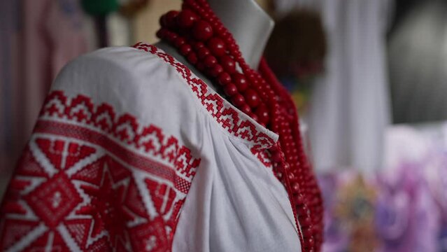 Close-up white dress with red traditional embroidery hanging on mannequin in craft shop. National Ukrainian costume with handmade accessories indoors. Slow motion