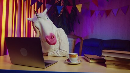 A woman in a unicorn mask is sitting on a chair at a table and typing on a laptop keyboard. This...