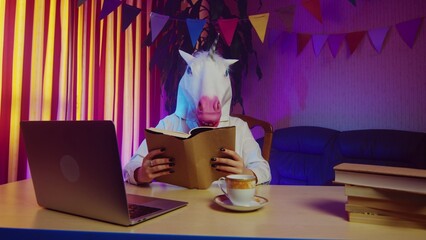 A woman in a unicorn mask is sitting on a chair at a table and reading a book. Multicolored...