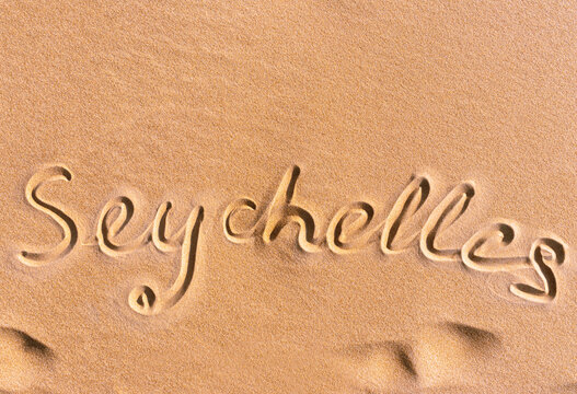 The text "Seychelles" is handwritten a on a golden sandy beach near the sea. Next to the text are waves of sand. Flat lay photo texture.
