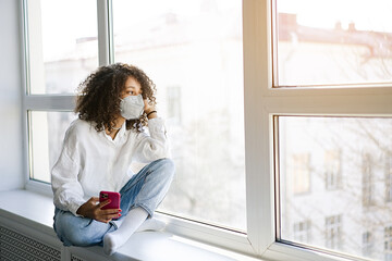 COVID-19 Pandemic Coronavirus. Young African american girl home isolation quarantine wearing face mask ffp2 protective for spreading of virus SARS-CoV-2. Woman chatting on a mobile phone at home 
