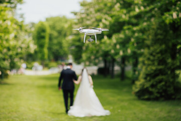 Photo of a drone filming wedding couple while they walk along in the park at wedding day.