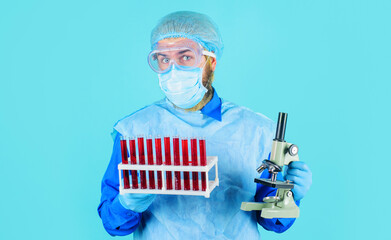 Doctor in protective suit, glasses and mask with blood sample tubes and microscope in laboratory.