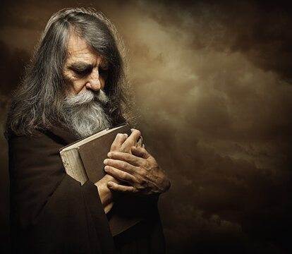 Praying Monk with Bible. Prophet holding Book. Old Man Portrait with Long Gray Beard in Black Cloak over Dark Mysterious Background