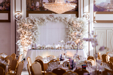 Front view of ornate wedding reception table, styled with golden candlesticks, white table cloths and dishes, placed on centre of hall on white background and arch with flowers. View of restaurant