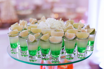 Close view of assorted shots with cocktail, which includes of green beverage, white tequila and piece of lime, located on glass plate  against blur background