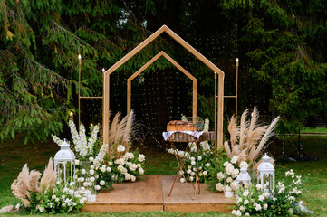 Front view of trendy boho wedding arch, which created of wood and decorated by lights and blooming bouquets of flowers, candles in glass and table with bread