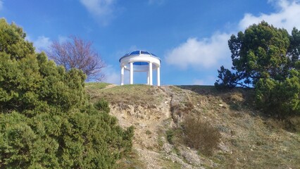 A rotunda with concrete columns stands on top of a hill. There are bushes and trees growing on the slope. The weather is sunny and the sky is blue with clouds