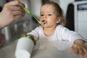 One baby close up on small caucasian girl and hand of her unknown mother feeding her at home in day small child with food around mouth making mess dirty while eating real people growing up copy space