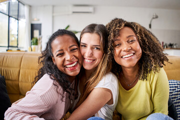 Obraz premium Three mixed race happy female friends hugging smiling - Funny women together celebrating sitting on the living room sofa looking at the camera selfie.