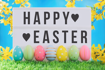 Bright positive spring Easter composition. Happy Easter text on lightbox against blue sky and yellow flowers. Multi-colored decorated eggs in a row on the grass.